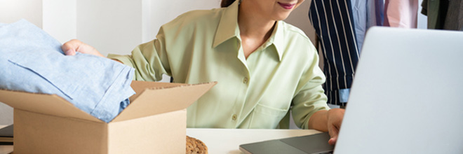 女性がパソコンを見ながら衣類をダンボールに梱包しようとしているイメージ画像