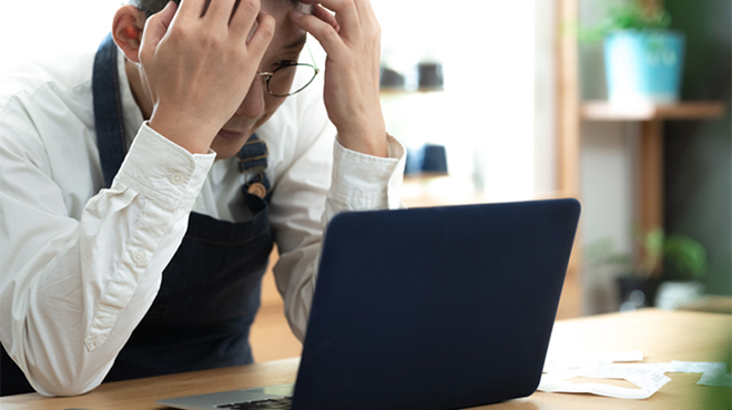 ノートパソコンの前で両手で頭を抱える男性のイメージ画像