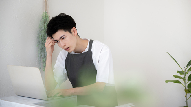 ノートパソコンの前で片手を頭にやって考えている様子の男性のイメージ画像