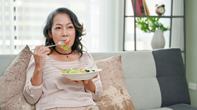 女性がソファーに座ってボウルに入ったサラダを食べているイメージ画像